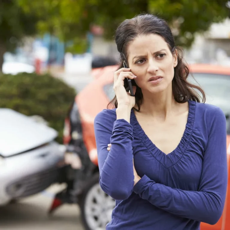 Concerned woman calling for help after a car accident, standing in front of two damaged vehicles on a suburban street.